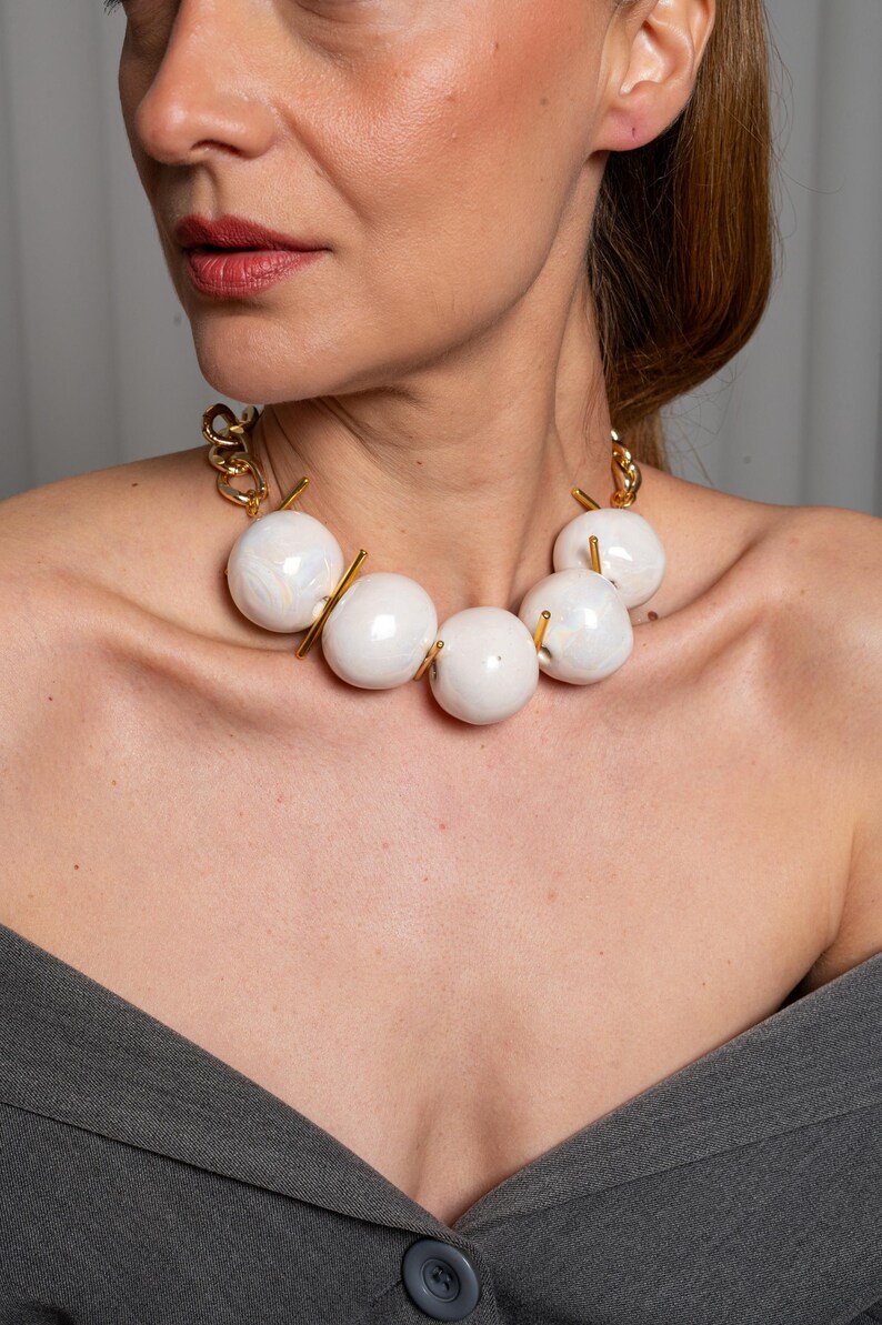 May include: A statement necklace featuring five large, smooth, off-white spherical beads with a subtle marbled finish. The beads are separated by thin, vertical gold-toned metal bars and connected to a thick gold-toned chain link necklace.