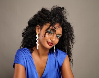 May include: Long white dangling earrings featuring a sharp, geometric zigzag shape. The earrings are attached to a small, circular gold-toned stud at the top. The design is minimalist and angular, hanging vertically from the earlobe in a clean, solid white finish.