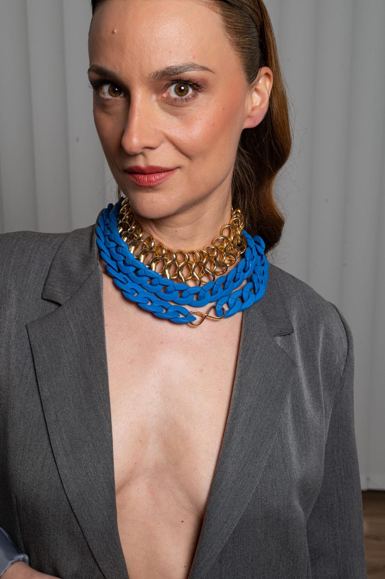 May include: A woman wearing a gray blazer with a layered necklace. The necklace is made of two blue plastic chains and a gold chain.
