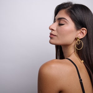 May include: A woman with long dark hair wearing gold hoop earrings with a circle and a curved line design. The earrings are dangling from her earlobes.