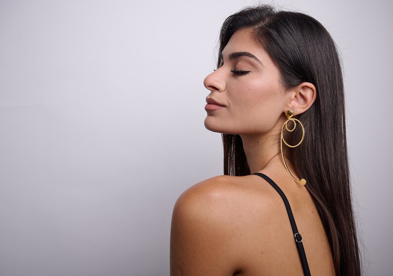 May include: A woman with long dark hair wearing gold hoop earrings with a circle and a curved line design. The earrings are dangling from her earlobes.