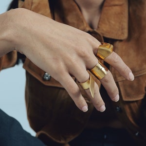 May include: Three gold-toned metal rings with a smooth, wide, and slightly irregular band design. Each ring features a distinct, open-ended shape that wraps around the finger, showcasing a polished metallic finish and a minimalist, structural aesthetic.