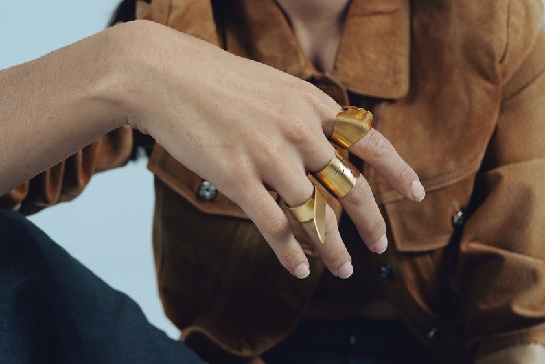 May include: Three gold-toned metal rings with a smooth, wide, and slightly irregular band design. Each ring features a distinct, open-ended shape that wraps around the finger, showcasing a polished metallic finish and a minimalist, structural aesthetic.