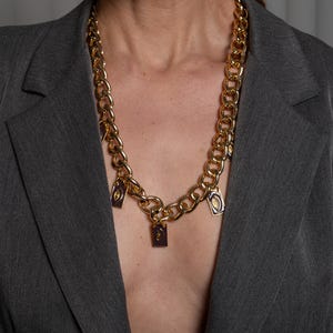 May include: A gold-toned chunky chain necklace featuring multiple rectangular purple enamel charms. Each charm displays a centered gold eye motif. The necklace is worn over a grey blazer, showcasing a bold link design with evenly spaced pendants.