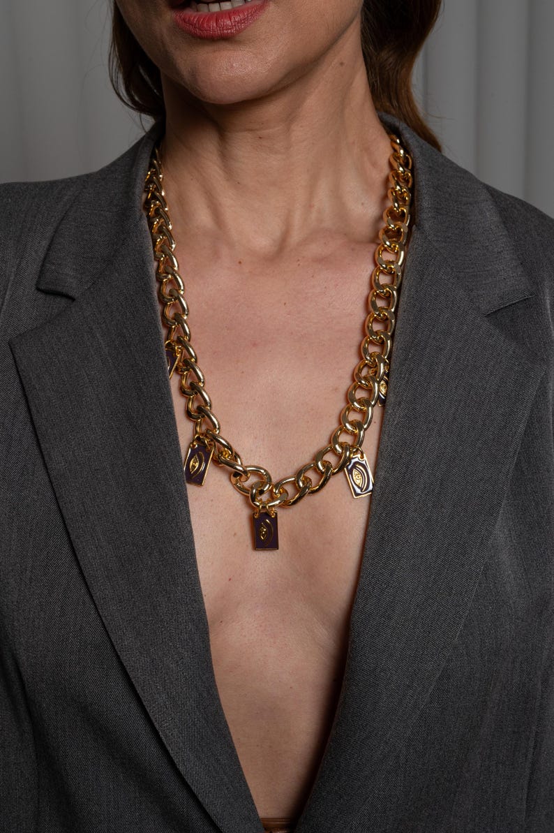 May include: A gold-toned chunky chain necklace featuring multiple rectangular purple enamel charms. Each charm displays a centered gold eye motif. The necklace is worn over a grey blazer, showcasing a bold link design with evenly spaced pendants.