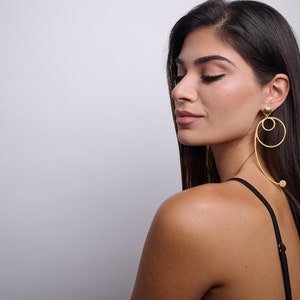 May include: A woman with long dark hair wearing a black tank top and gold hoop earrings with a dangling ball accent.