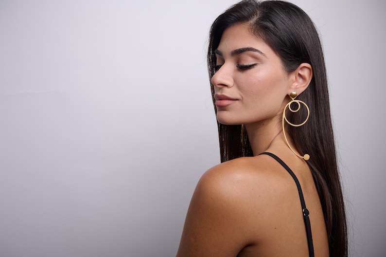 May include: A woman with long dark hair wearing a black tank top and gold hoop earrings with a dangling ball accent.