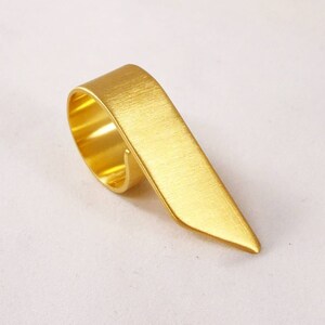 May include: A gold-colored ring with a brushed metal finish. The band features a continuous loop design that extends into a long, flat, pointed triangular extension that tapers to a sharp tip, creating a distinct geometric silhouette.