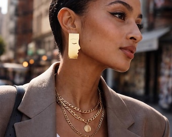 May include: A gold-toned rectangular earring featuring a minimalist line-art profile of a face with closed eyes. The earring is paired with a layered gold-toned chain necklace that includes a small circular pendant hanging from the center chain.