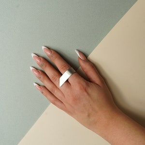 May include: A silver ring with a wide, flat band. The ring is worn on the index finger of a hand with white French manicured nails.