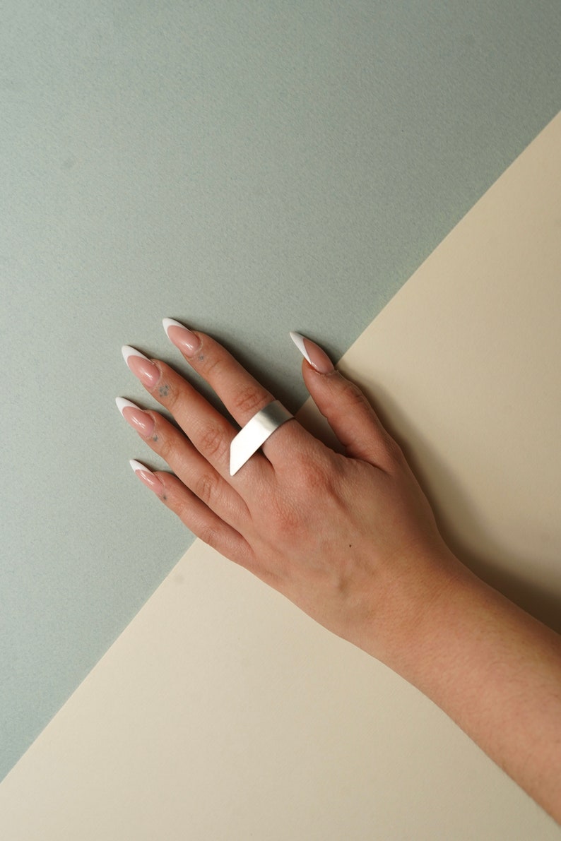 May include: A wide, matte silver ring with an angular, slanted top edge. The band features a smooth, metallic finish and a geometric design that wraps around the finger, creating a bold and clean aesthetic for this piece of hand jewelry.