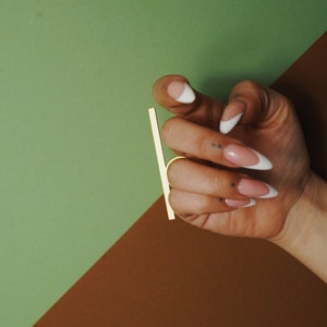 May include: A hand with long, almond-shaped nails featuring a classic French manicure with white tips and a nude base. The hand is positioned next to a thin, vertical gold-colored metal bar, showcasing a clean and polished aesthetic for nail art.
