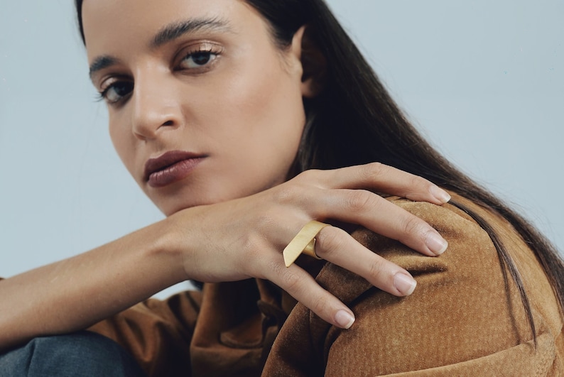 May include: A gold-toned ring featuring a wide, flat, rectangular bar design that sits horizontally across the finger. The metal has a smooth, matte finish, and the band is circular, providing a clean and simple aesthetic for the hand.
