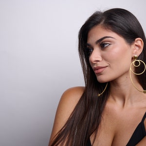 May include: A woman with long dark hair wearing a black vest top and gold hoop earrings with a geometric design.
