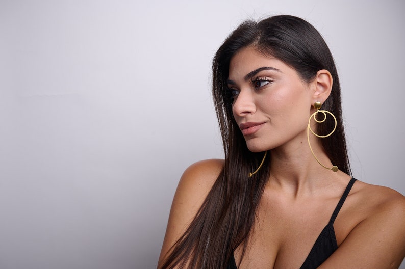 May include: A woman with long dark hair wearing a black vest top and gold hoop earrings with a geometric design.