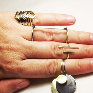 May include: A hand wearing a set of gold and silver rings. The rings are stacked on the index finger and include a geometric design, a circle with a hole, and a rectangle. The middle finger has a silver ring with a gold bar attached. The pinky finger has a silver ring with a gold disc attached.