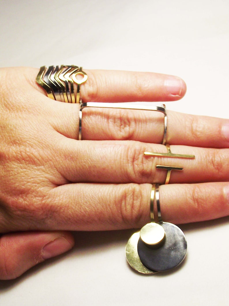 May include: A hand wearing a set of gold and silver rings. The rings are stacked on the index finger and include a geometric design, a circle with a hole, and a rectangle. The middle finger has a silver ring with a gold bar attached. The pinky finger has a silver ring with a gold disc attached.