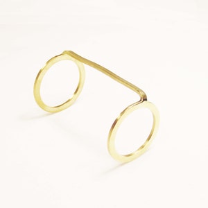 May include: A gold-coloured metal ring with two circular openings. The ring is made from a single piece of metal that is bent into a U-shape.