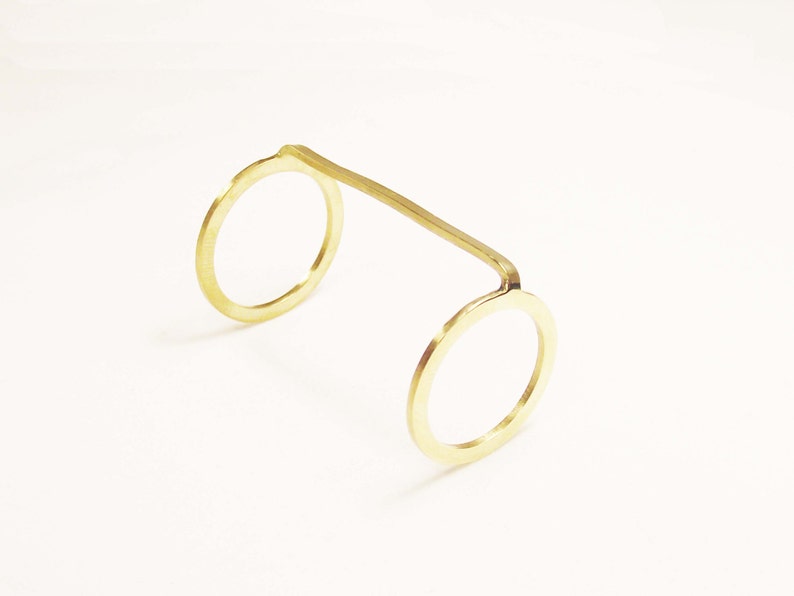 May include: A gold-coloured metal ring with two circular openings. The ring is made from a single piece of metal that is bent into a U-shape.
