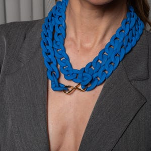 May include: A woman wearing a gray blazer with a double-layered blue plastic chain necklace with a gold infinity charm.
