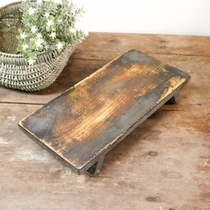 May include: A rectangular, rustic wooden serving tray with a distressed finish. The tray is supported by short legs and has a dark, weathered appearance with hints of brown and gray. A woven basket with greenery is in the background.