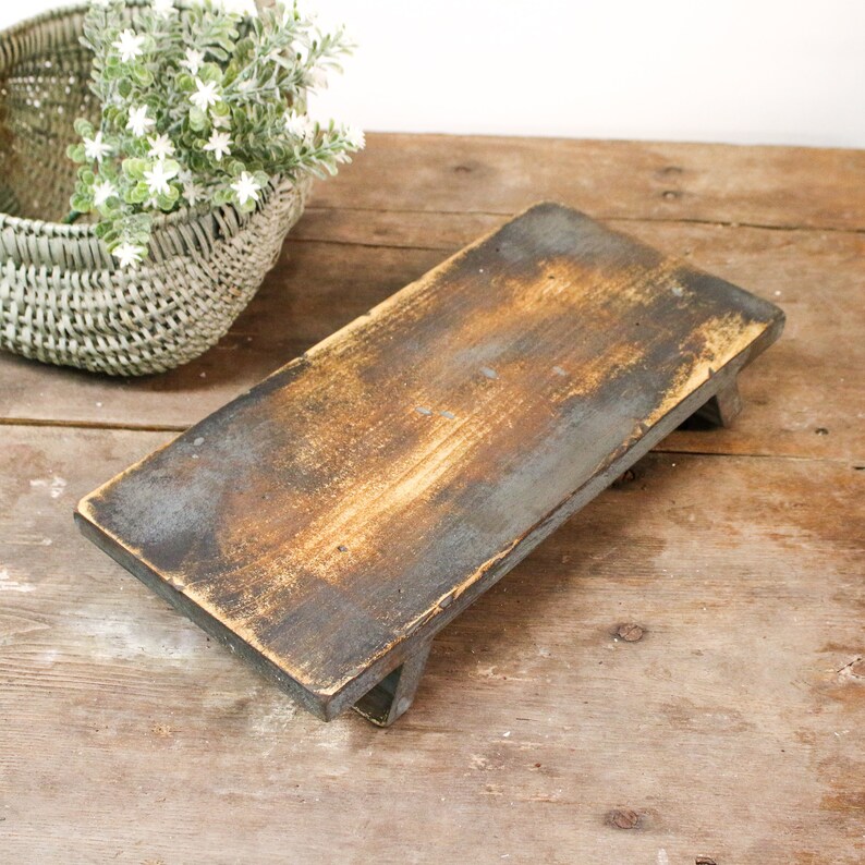 May include: A rectangular, rustic wooden serving tray with a distressed finish. The tray is supported by short legs and has a dark, weathered appearance with hints of brown and gray. A woven basket with greenery is in the background.