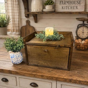 May include: A rectangular wooden bread box with a hinged front door and a small round knob. A yellow pillar candle sits on top of the box, surrounded by a ring of small green artificial leaves. The wood has a dark brown, stained finish.