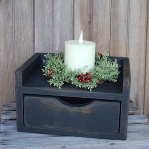 May include: A black wooden nightstand with a drawer. A cream-colored candle sits atop the nightstand, surrounded by a green wreath with red berries. The nightstand is on a wooden surface.
