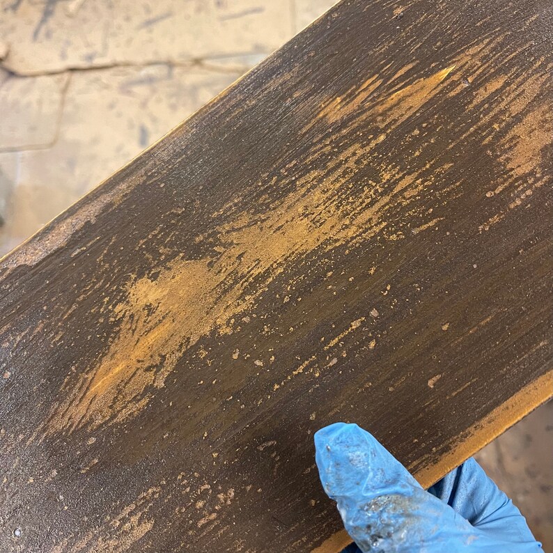 May include: Close-up of a distressed wooden plank with a dark brown base and golden-colored accents. The wood grain is visible, and the surface has a textured, weathered appearance. The plank is angled, showing its edge.