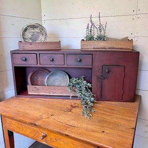 May include: A rustic, burgundy-colored cabinet with three drawers, a small door, and an open shelf. It features a wooden table with a drawer. Decorative items include wooden trays and bowls, and a lavender plant.