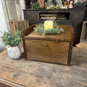 May include: A rustic, wooden bread box with a hinged door and a small wooden knob. The box is stained a warm brown and has a small ledge on top. A faux candle and greenery sit on top of the box. A blue and white floral vase is to the left.