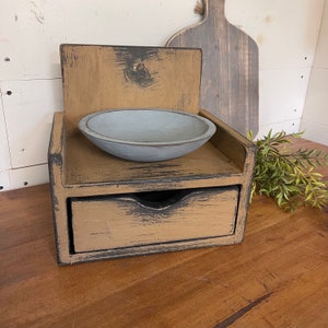 May include: A distressed wooden organizer with a small drawer and a shelf holding a light blue bowl. The organizer has a tan finish with black accents. A wooden cutting board and greenery are in the background.