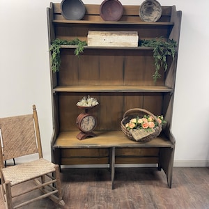 May include: A rustic, wooden hutch with three shelves, holding decorative bowls, a small box, and greenery. A vintage scale and a basket of flowers sit on the bottom shelf. A woven chair is to the left. The hutch is painted in shades of brown and gray.