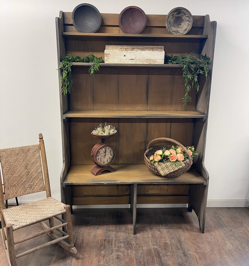 May include: A rustic, wooden hutch with three shelves, holding decorative bowls, a small box, and greenery. A vintage scale and a basket of flowers sit on the bottom shelf. A woven chair is to the left. The hutch is painted in shades of brown and gray.