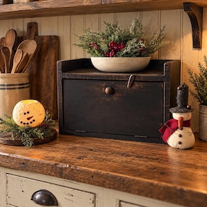 May include: A dark brown wooden bread box with a drop-down door and a small knob, topped with a white bowl filled with green pine branches and red berries. A snowman figurine with a black hat and red scarf sits beside the bread box.