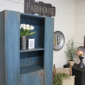 May include: A blue farmhouse-style cabinet with open shelving and a lower door. The cabinet is adorned with a wooden sign that reads "FARMHOUSE." Inside, a metal vase holds white tulips, and a small sign says "love grows here."