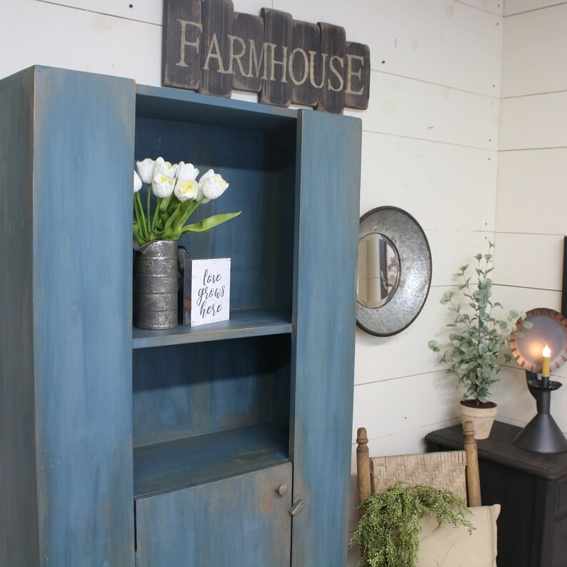 May include: A blue farmhouse-style cabinet with open shelving and a lower door. The cabinet is adorned with a wooden sign that reads "FARMHOUSE." Inside, a metal vase holds white tulips, and a small sign says "love grows here."