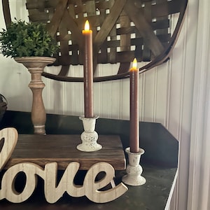 May include: Two brown pillar candles with a warm glow are displayed on white distressed metal candle holders. The candles are on a wooden surface with the word "love" written in white on the surface.