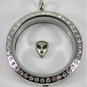 May include: A silver locket with a clear glass insert and a silver alien charm inside. The locket is surrounded by a row of clear crystals.