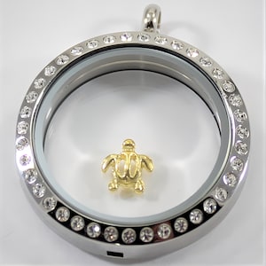 May include: A silver locket with a clear glass insert and a gold turtle charm. The locket is surrounded by a row of clear rhinestones.