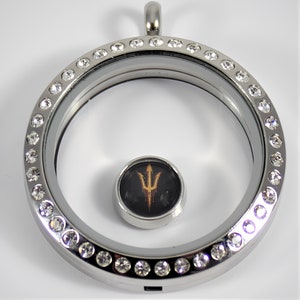 May include: Silver locket with a clear glass insert and a small round charm featuring a gold trident logo on a black background. The locket is adorned with a row of clear crystals around the edge.