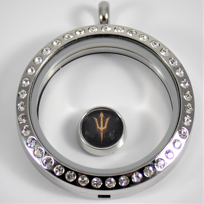 May include: Silver locket with a clear glass insert and a small round charm featuring a gold trident logo on a black background. The locket is adorned with a row of clear crystals around the edge.