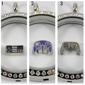 May include: Three silver locket charms with different video game controller designs. The first charm features a black and red NES controller, the second charm features a purple GameCube controller, and the third charm features a silver Xbox controller with colorful buttons.