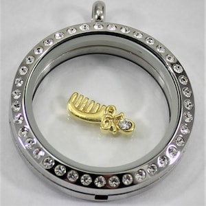 May include: A silver-colored, round locket with a clear center, encrusted with small, clear crystals around the edge. Inside, a gold-colored charm shaped like a comb and a baby shoe with a crystal accent.