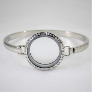 May include: A silver bangle bracelet with a round locket featuring a clear glass insert and a border of small clear rhinestones.