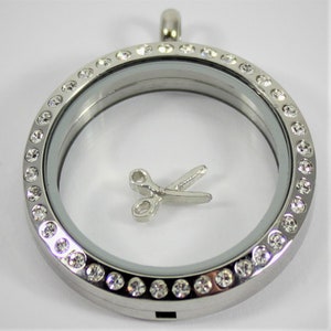 May include: A silver locket with a clear glass insert and a silver pair of scissors charm. The locket is surrounded by a row of clear rhinestones.