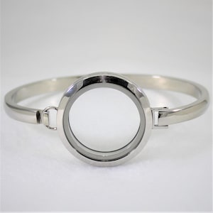 May include: A silver bangle bracelet with a round, clear glass locket attached. The locket is open and ready to hold a small photo or other memento.