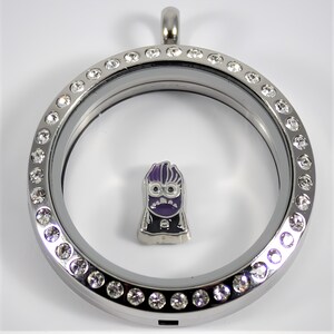 May include: A silver locket with a clear glass insert and a silver charm inside. The locket is decorated with a circle of clear rhinestones. The charm is a purple cartoon character with a black outline.