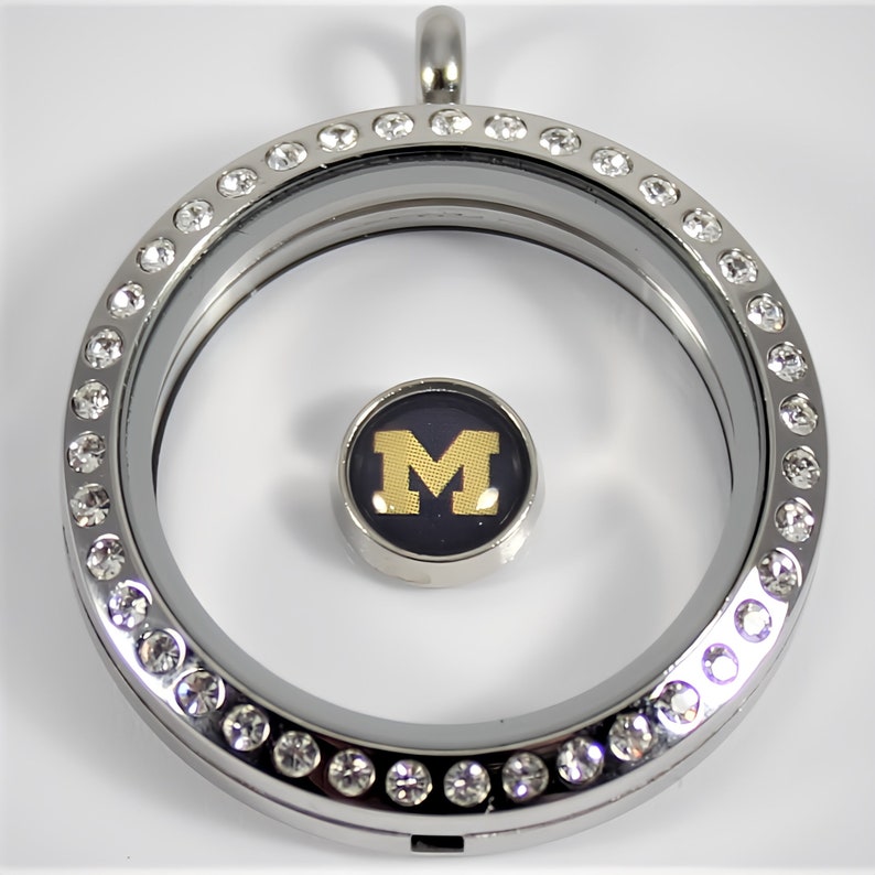 May include: Silver locket necklace with a clear glass insert and a crystal-encrusted border. The locket contains a small charm with a black background and a gold "M" in the center.