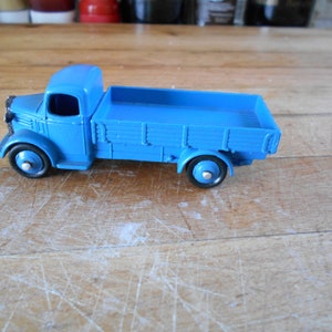 May include: A blue toy truck with a flatbed and wooden sides. The truck has a cab with a windscreen and two wheels on each side.